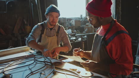 blacksmith measuring wrought iron as colleague writing results in workshop