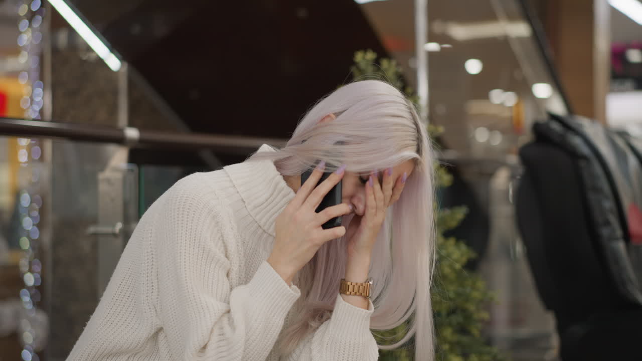 mall wanderer seated on bench removes phone from ear while listening intently, wearing white sweater, jeans and boots with black purse beside her, glass railing and decorative lights behind