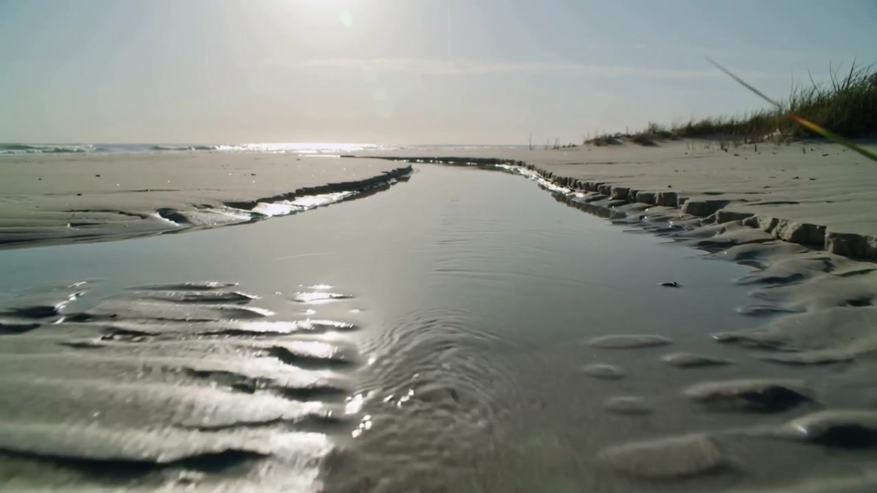 Abfließendes Rinnsal nach Regenwasserabfluss, das über den Strand fließt und sich zu einem ruhigen Becken mit Dünengras verbreitert.