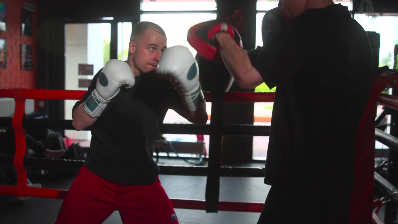 Jovem boxeador masculino pratica trabalho de manopla dentro do ringue de boxe com o treinador, mantendo-se baixo em guarda defensiva, luvas brancas perto do queixo, cordas vermelhas, forte contraluz da janela, contraste granulado e energia focada
