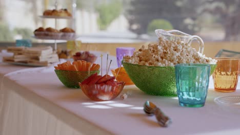 approaching to a table with sweets for a party in summer, with a tray with croissants, sandwich, popcorn, crudités, coloured glasses and white tablecloth moving blowed by the wind