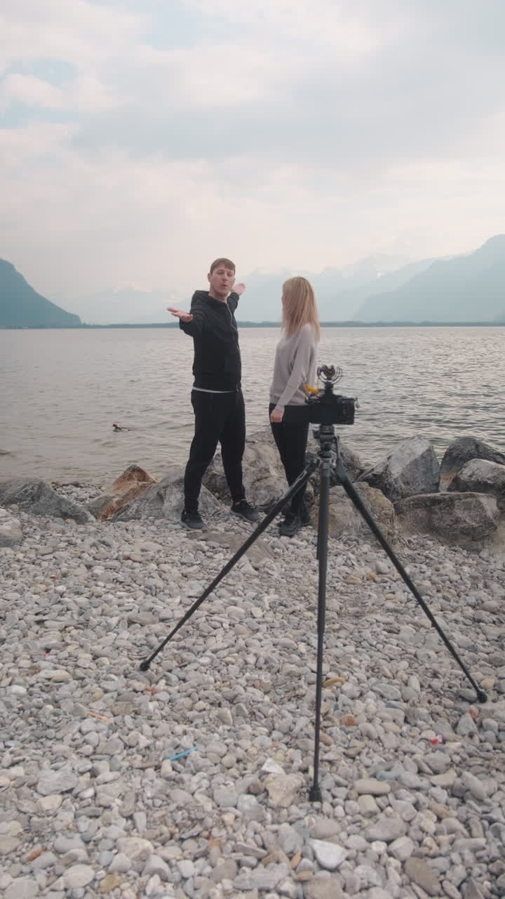 Vertical: pareja caucásica vlogueando frente a un trípode con cámara y el Lago Lemán en Montreux, Suiza, toma estática