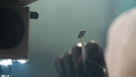 una vista de cerca de la mano con guantes de un técnico sosteniendo un pequeño componente electrónico con pinzas bajo un microscopio