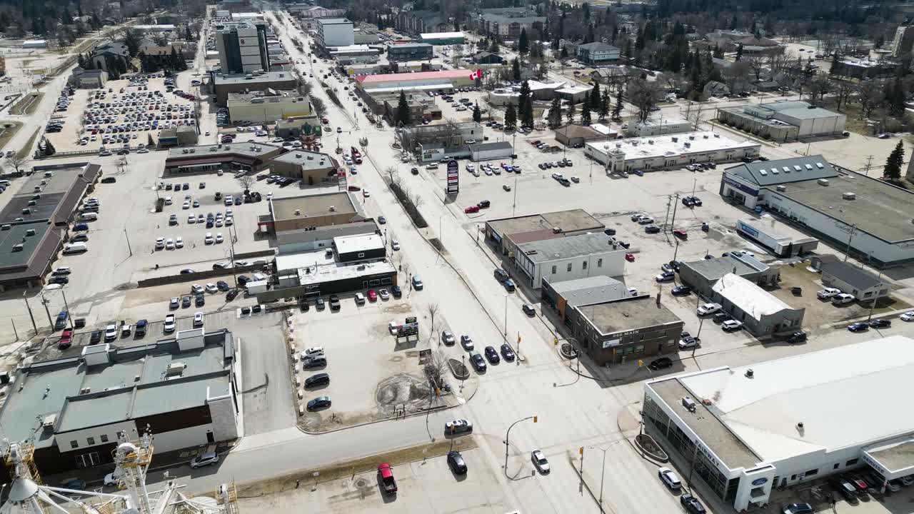 Ripresa aerea lenta con drone che guarda dall'alto una piccola città di giorno con auto che percorrono le strade e attività commerciali visibili, parcheggi e forte luce solare che crea ombre