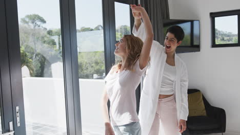 feliz pareja de lesbianas caucásicas bailando junto a la ventana y abrazándose en la casa soleada