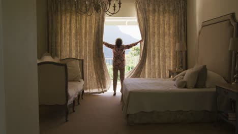 rear view of caucasian senior woman opening window curtains at home