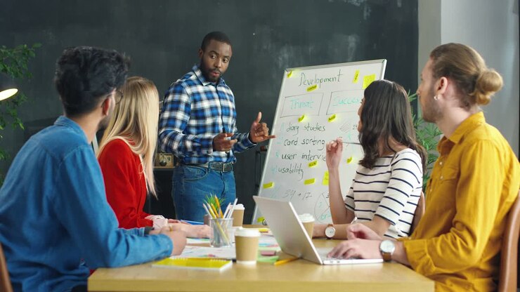 Homme expliquant une stratégie commerciale avec un tableau noir au bureau, puis ses collègues applaudissent