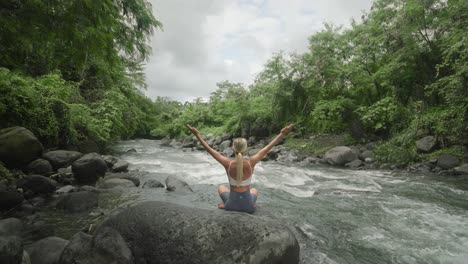 rear view of blond woman raising arms in lotus pose on rock next to river, yoga