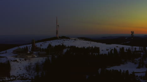 Toma-Aérea-Al-Amanecer-Que-Asciende-Sobre-Una-Colina-Cubierta-De-Nieve-Que-Muestra-La-Torre-De-Radiodifusión,-La-Turbina-Eólica-Y-Los-Edificios