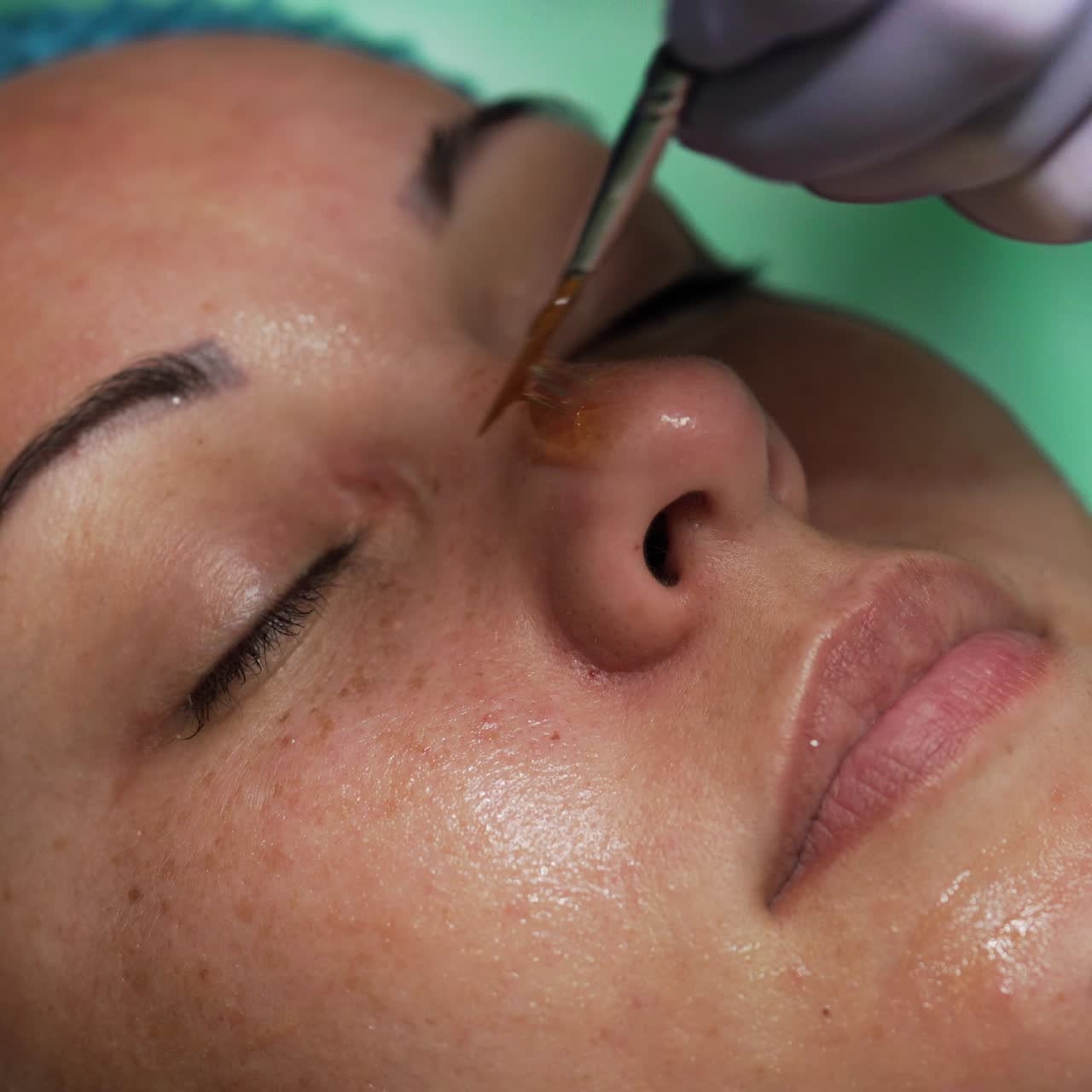 Face of woman is covered with yellowish gel. Beauty specialist uses brush to apply the remedy. Extreme close up