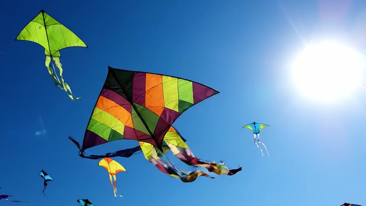 Una Vivace Esibizione di Aquiloni Colorati che Volano in un Cielo Blu Chiaro, Illuminati dalla Luminosa Luce del Sole, Creando una Scena Gioiosa di Libertà e Movimento Sopra il Suolo