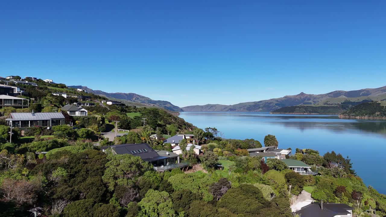 Aerial views of Akaroa's serene coastline, showcasing lush greenery, calm waters, and residential areas under clear blue skies