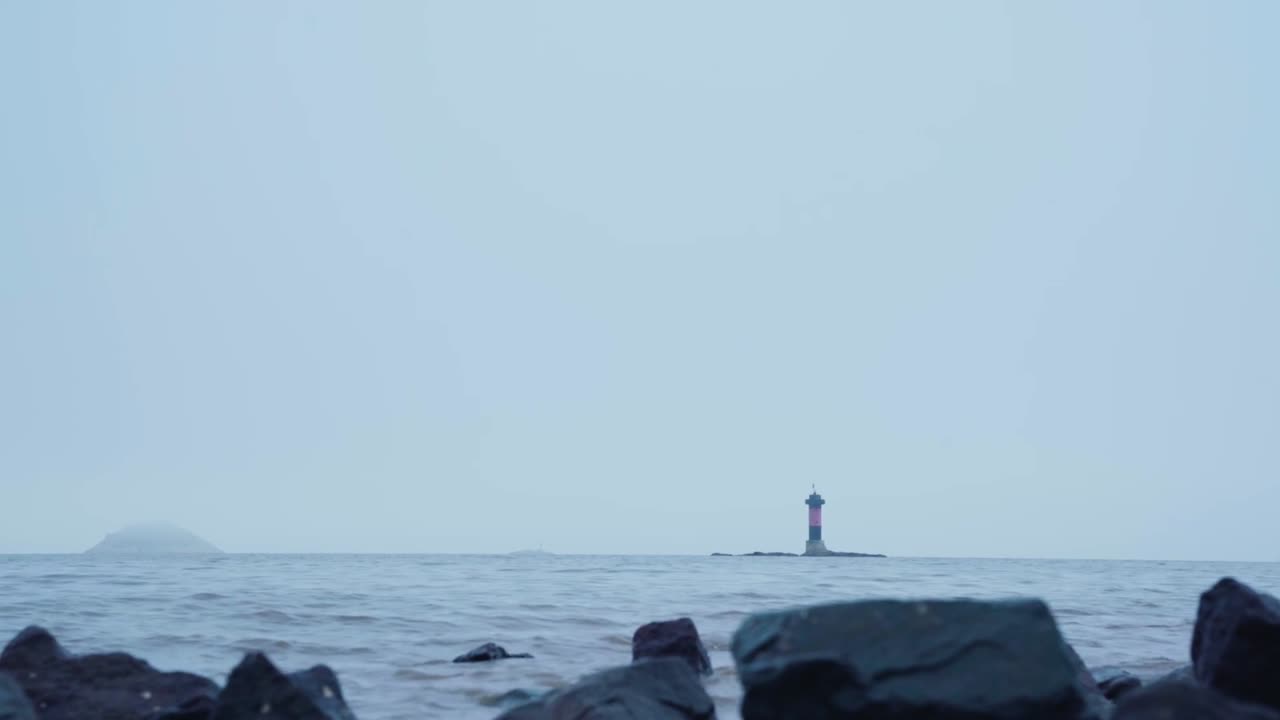 Red lantern tower of Longstone Lighthouse stands isolated on rocky outcrop in overcast sea, subtle island visible on horizon, tranquil and remote Farne Islands coastal atmosphere