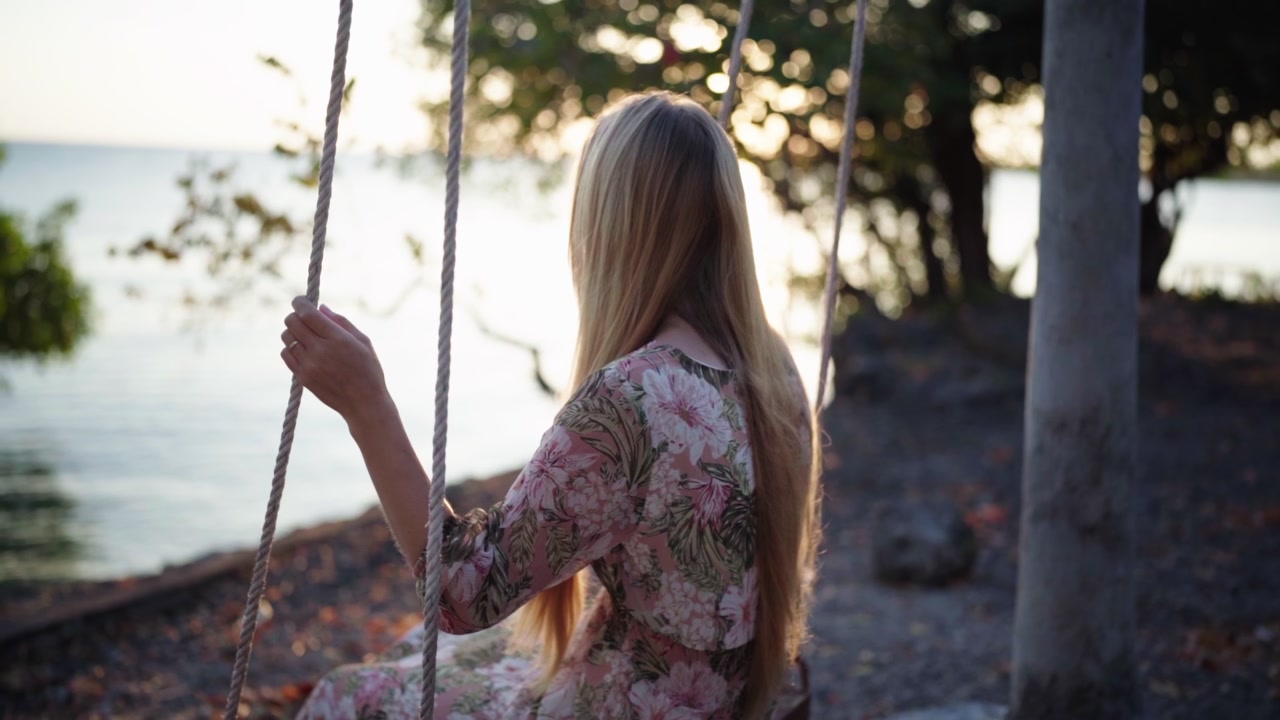 A woman gently swinging by the seaside with soft morning sunlight creating a beautiful backlight. Surrounded by green trees and coastal breeze, capturing a peaceful tropical lifestyle moment.