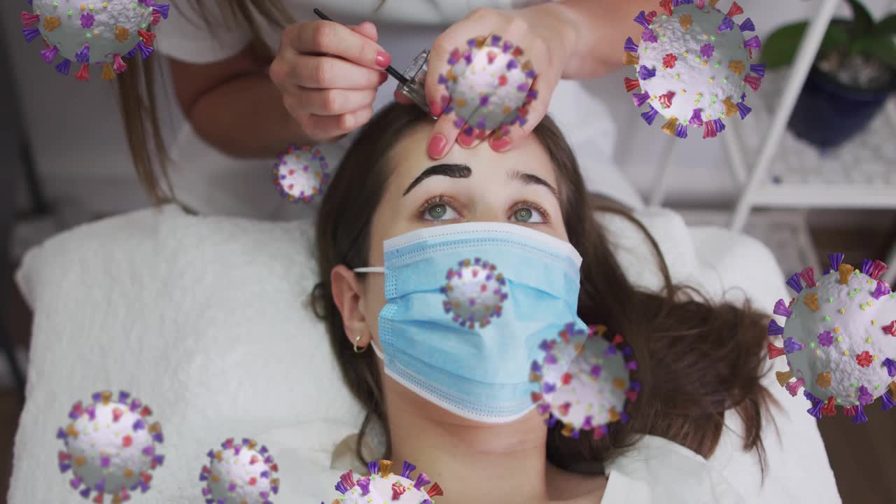 Mentre intinge il pennello nella tinta, la cosmetologa nel salone pettina le sopracciglia della cliente mentre fluttuano icone di virus