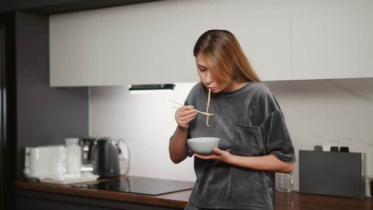Young lady with long hair resting against kitchen cabinet eating noodles with chopsticks, wearing casual gray outfit, bright modern kitchen interior with soft lighting