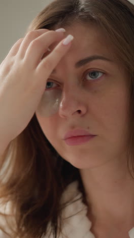 mujer de cabello oscuro hace tratamientos de belleza de pie en un baño espacioso. la dama cuida la piel facial usando parches debajo de los ojos después de bañarse