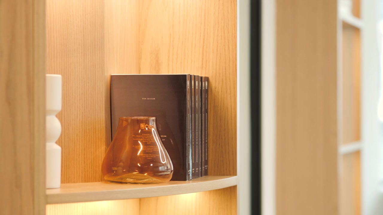 Detail of light wood furniture with warm lighting inside: dark-covered books, an amber glass vase and a minimalist white decorative piece are on display