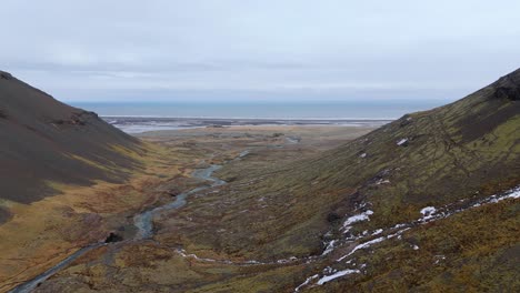 Glacier-river-flowing-from-mountain-valley-into-vast-plains,-gray-sky