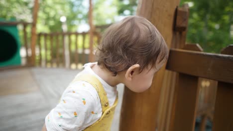 adorable niñita apoyada en la columna de madera y mirando tirar la cerca de madera por curiosidad en el patio de recreo al aire libre de los niños durante el día, primer plano en cámara lenta