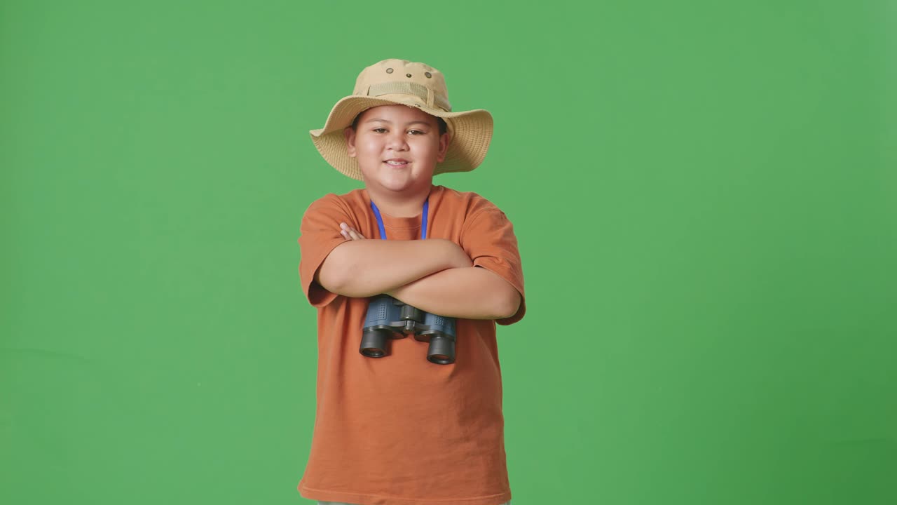 모자와 안경을 입은 아시아 관광 소년은 녹색 스크린 배경에 서서 팔을 고 카메라에 미소 짓고 있습니다. 소년 연구자는 여행 관광 모험 개념을 조사합니다.