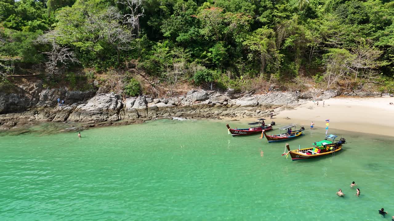 las imágenes de drones capturan vibrantes barcos de cola larga y nadadores en una aislada playa de phuket, mostrando una exuberante vegetación y aguas turquesas.