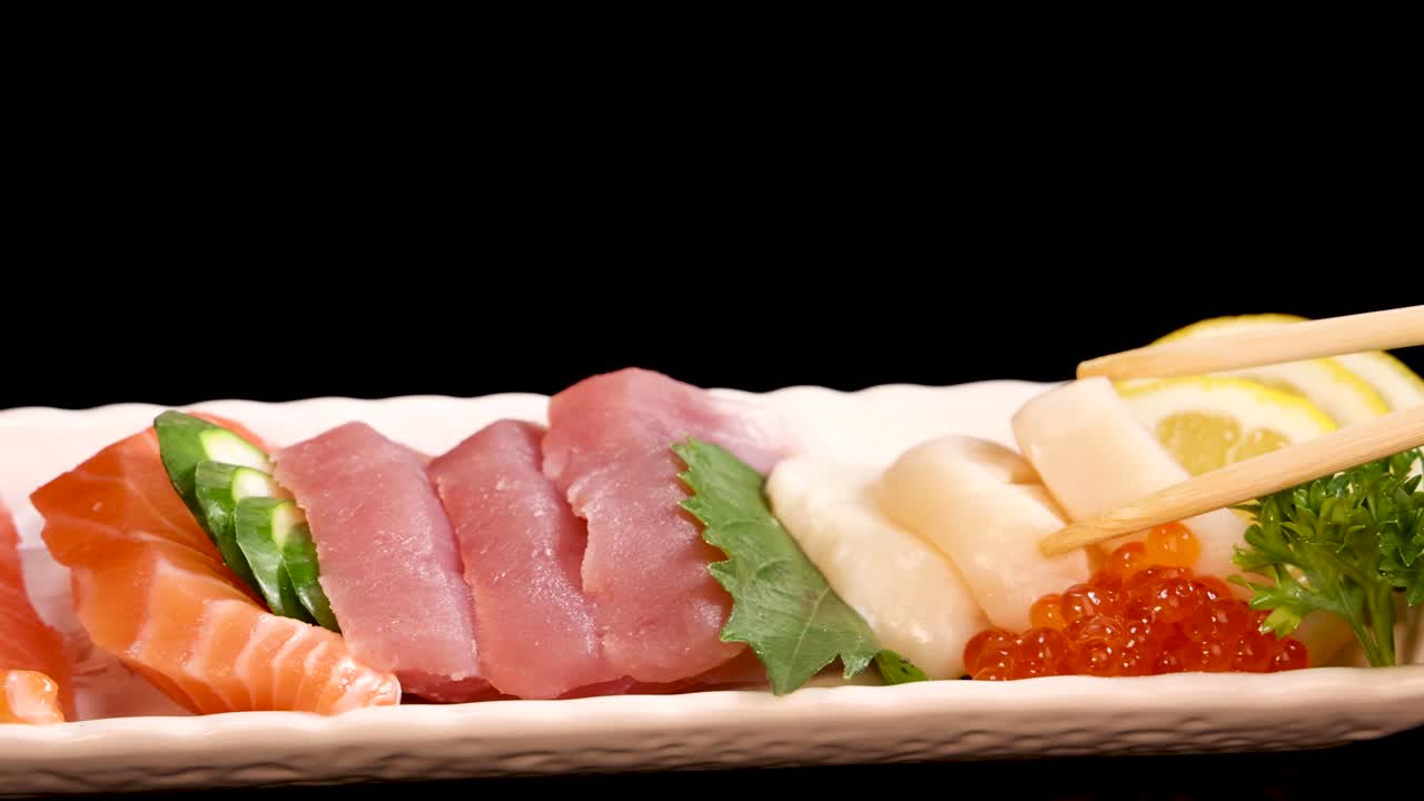 Chopsticks select pieces of salmon sashimi and scallop sushi from a neatly arranged platter under studio lighting, highlighting fresh seafood and vibrant garnishes