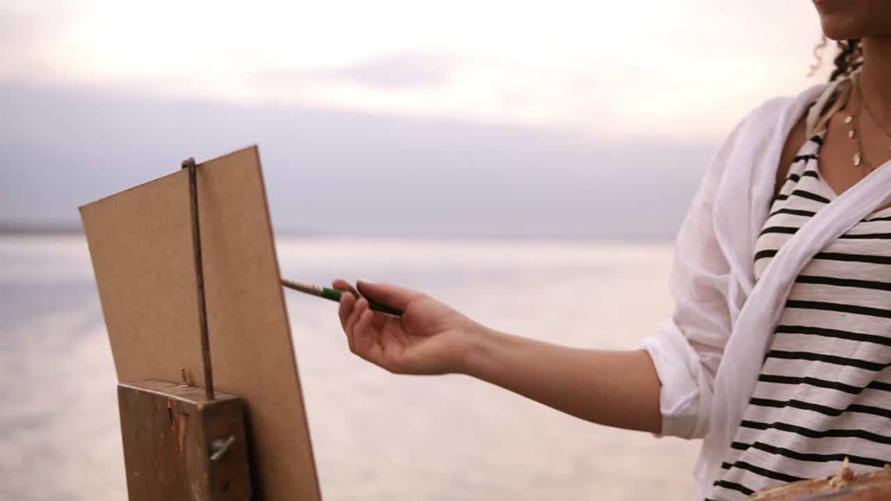 Close up of an artist's art process. An easel and palette. Girl is putting some color paints on canvas. Blurred lake surround on the background