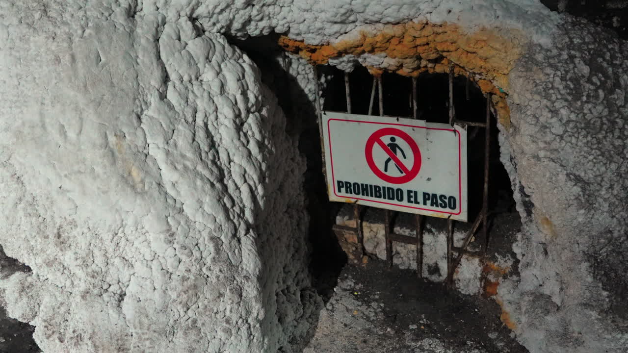 Intriguing close-up of a 'Prohibido el Paso' sign at a cave entrance, bathed in dramatic shadows. The textured rock and rusty bars create a mysterious, forbidding atmosphere
