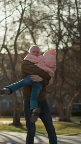un hombre con una chaqueta marrón y pantalones vaqueros azules llevando y girando alegremente a una niña con una gorra rosada y una chaqueta en un parque iluminado por el sol. un momento lúdico y conmovedor de unión padre-hija