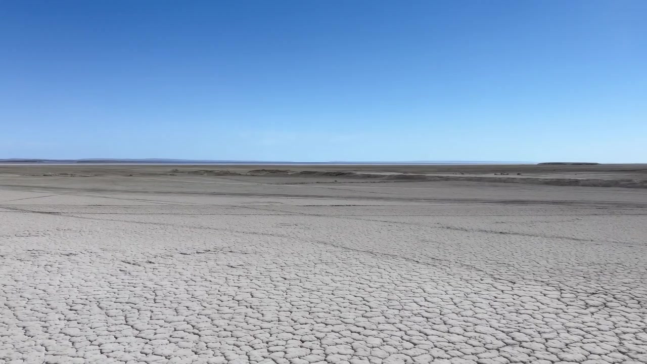 Elevated panoramic view showing cracked dry lakebed spreading from dense polygonal mud plates in foreground into smoother fractured ground toward flat horizon beneath clear pale blue sky, creating
