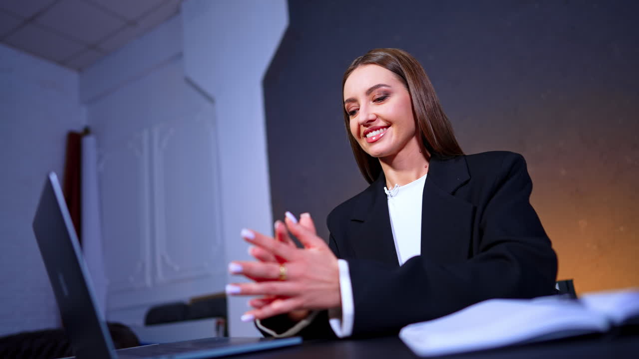 Close up portrait of female presenter wearing black blazer and microphone. Woman in formal attire sitting at desk with laptop looking away from camera in professional television studio