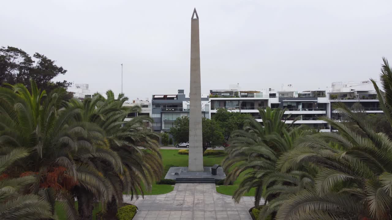 높은 오벨리스크 기념물과 많은 나무와 푸른 풀이 있는 공원에서 드론이 이륙하고 고도를 오르는 4k 비디오, 아파트 건물 뒤에