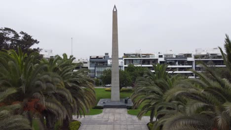 4k-video-of-a-drone-lifting-off-and-climbing-altitude-in-a-park-with-a-tall-obelisc-monument,-and-many-trees-and-green-grass,-behind-it-apartment-buildings