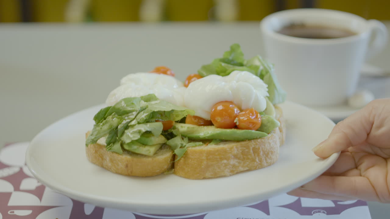 Close-up of a plate with rustic bread toast topped with avocado, lettuce, cherry tomatoes and poached eggs, next to a cup of hot coffee