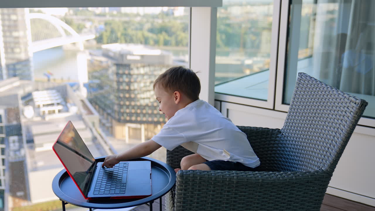 Happy active baby boy jumps in the armchair. Kid looks at laptop smiling and presses the key on the keyboard