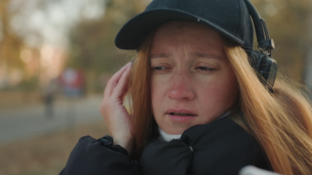 Gros plan sur une femme rousse avec un casque audio réagissant à un voleur à proximité dans un parc sous une lumière automnale, doudoune noire, casquette et écharpe, expression anxieuse, main à l'oreille, regard scrutateur, portrait de rue spontané.