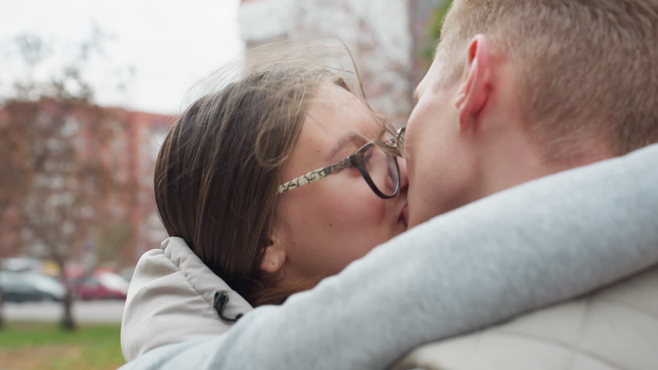 Close up of husband and wife kissing affectionately outdoors while holding each other warmly, sharing romantic moment in casual matching jackets with trees and blurred city background
