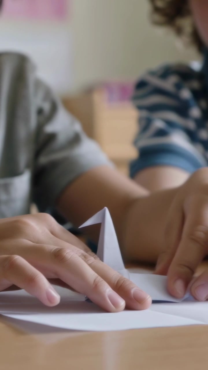 Video verticale: Raccogliendo carta alla scrivania, bambini in maglietta grigia e polo a righe che piegano la carta per costruire una tenda per la lezione.