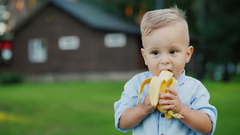 Fröhliches-1-jähriges-Baby-Isst-Eine-Banane,-Es-Steht-Auf-Der-Rückseite-Des-Hauses-4k-Video