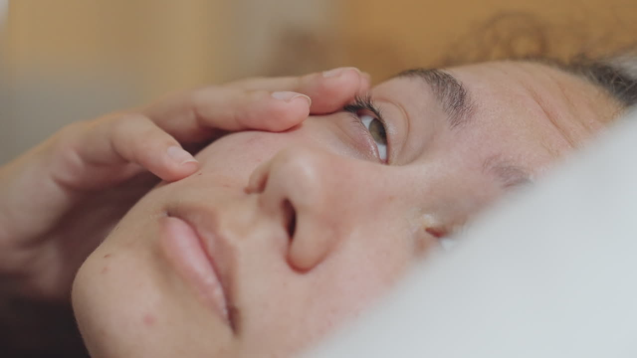 Mujer somnolienta despertando en la cama del hotel, primer plano de una huésped contemplativa en una almohada blanca con suave luz matutina, movimiento suave de ojos y roce sutil de manos, ambiente tranquilo e íntimo, grano cinematográfico.