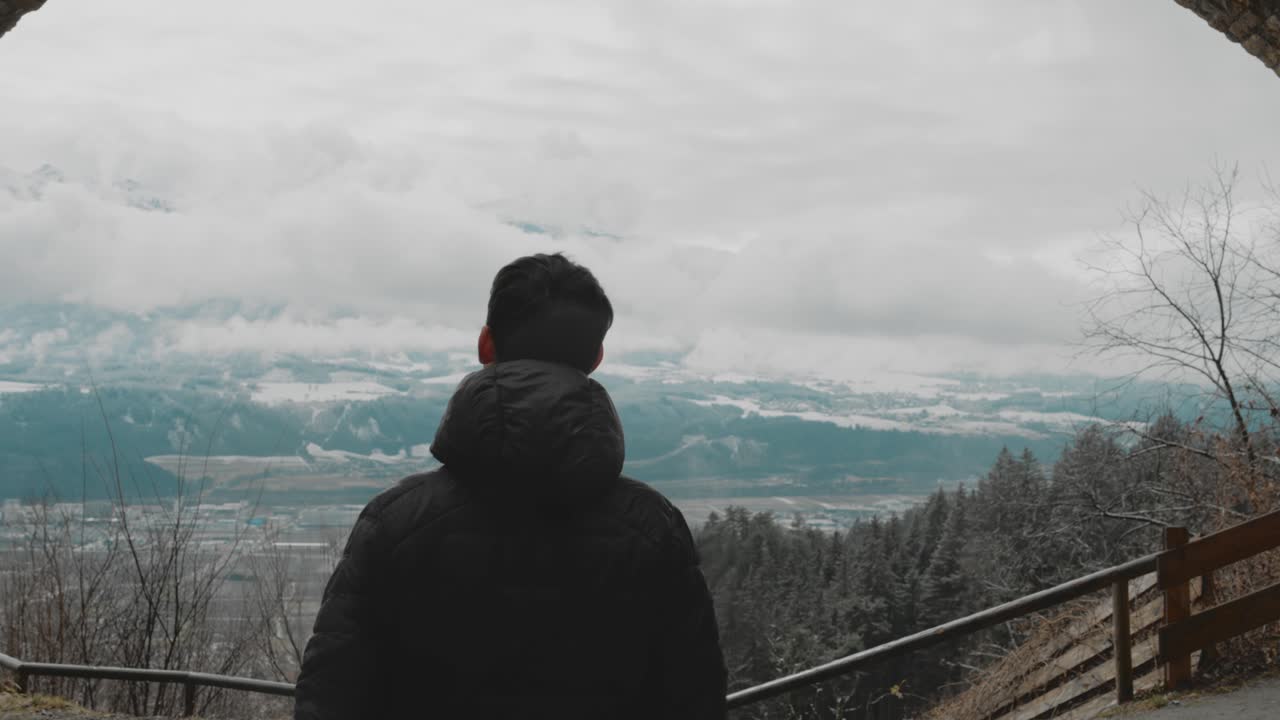 짧은 어두운 머리카락을 가진 젊은 남자가 구름과 눈 인 산과 계곡의 숲과 함께 먼 곳을 바라보는 고대 폐허 아크를 걸어가고 있습니다.