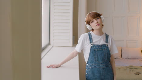 Girl-Standing-Near-A-Window-Wearing-Headphones,-Then-Smiling-And-Looking-To-The-Camera