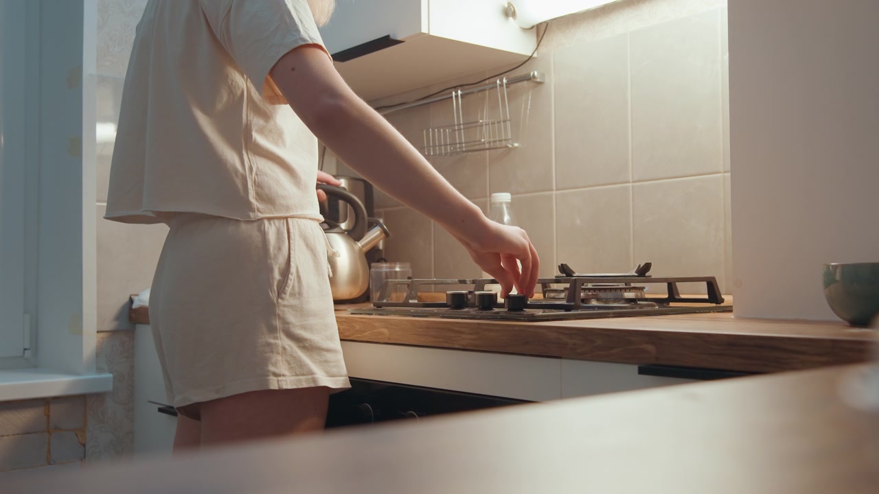 Cucina mattutina delicata, cuoco/a casalingo/a che regola il fornello, scena di colazione tranquilla con bollitore sul fornello a gas, piano di lavoro in legno, tazza in ceramica, calda luce solare attraverso la finestra, comfort dell'abbigliamento da casa e vapore delicato che sale.