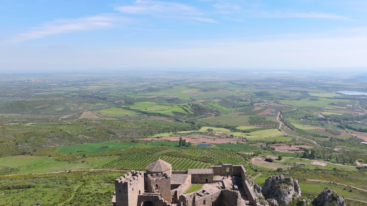 Reverse drone shot revealing a medieval castle atop a rocky outcrop, surrounded by forest and vibrant agricultural fields under a clear blue sky. Scenic historical architecture in rural Spain