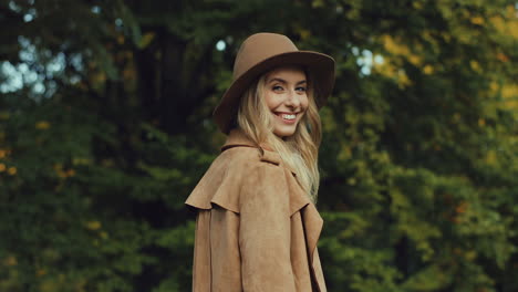 Rear-view-of-Caucasian-blonde-young-woman-in-a-hat-and-coat-in-the-park-in-autumn,-then-turns-and-looks-at-the-camera