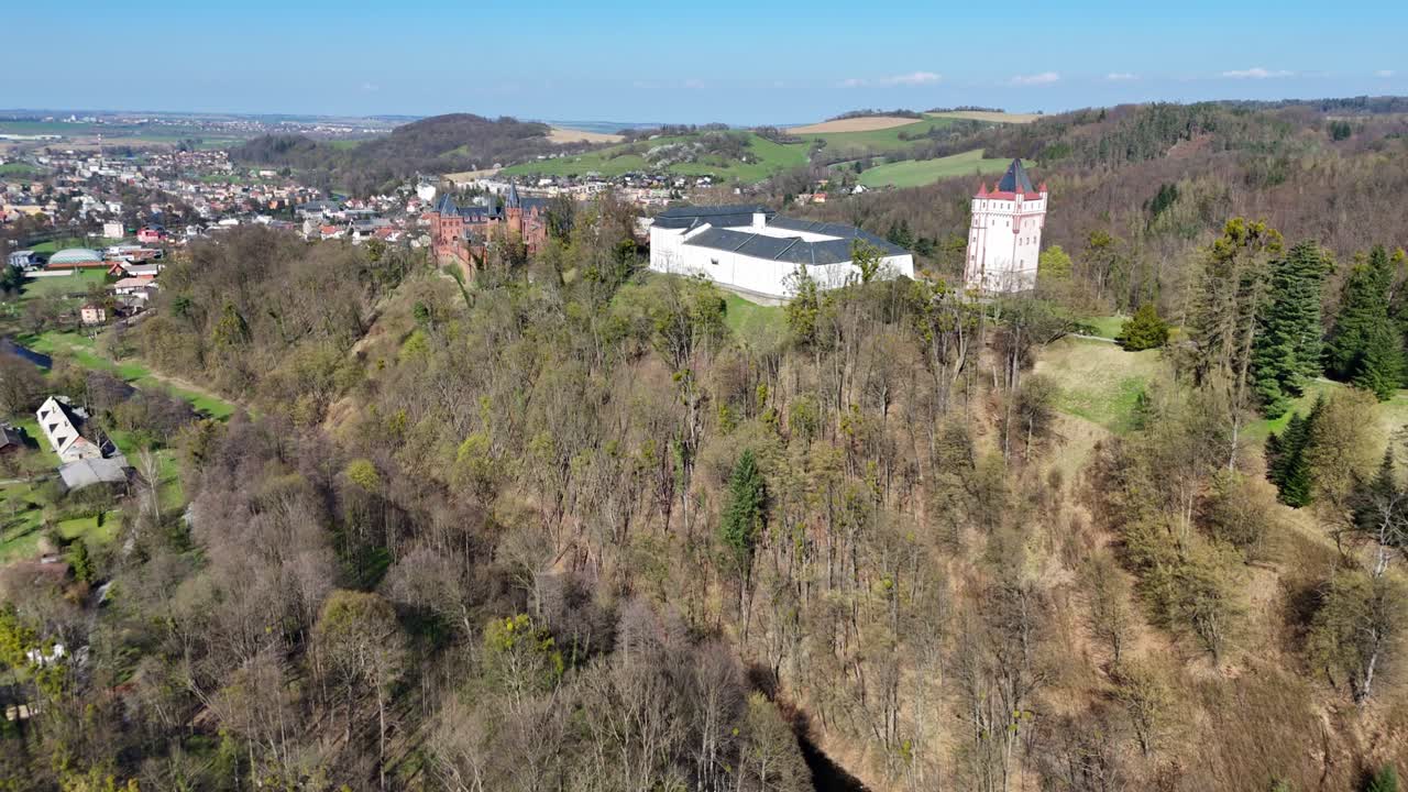 Fremoverbevegelse mens dronen sakte synker og nærmer seg Hradec nad Moravicí slott i Tsjekkia, med skogkledd åsside, nærliggende by og vidstrakt utsikt over landskapet.