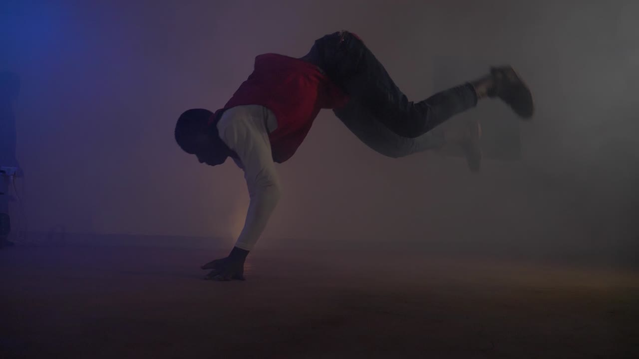 Slow motion shot of African youth in red shirt doing a break-dancing move jumping from his hands to feet in colorful disco lights and smoke