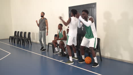 multiracial male basketball players supporting game, celebrating and raising hands with coach in gym