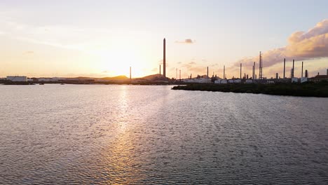 Drone-push-in-to-flare-stacks-over-water-at-sunset,-golden-hour-sky